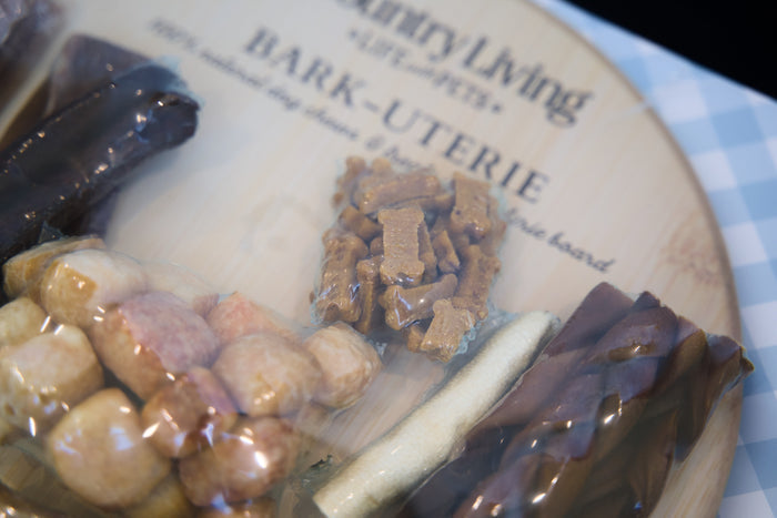Assorted dog treats on a branded wooden board with a checkered tablecloth.|Friandises pour chiens assorties sur une planche en bois de la marque, posée sur une nappe à carreaux.