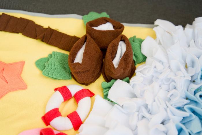 Close-up of fabric palm tree and lifebuoy designs on a beach-themed pet snuffle mat.|Gros plan sur des motifs de palmiers et de bouée de sauvetage en tissu sur un tapis de fouille pour animaux à thème plage.