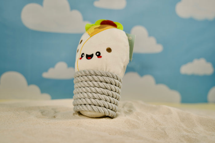 Dog toy shaped like a burrito resting on a sandy beach.|Jouet pour chien en forme de burrito posé sur une plage de sable.