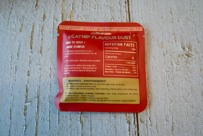 A red packet of Catnip Flavour Dust cat food placed on a weathered white wooden surface.|Un sachet rouge de poudre aromatisée à l'herbe à chat pour félins, posé sur une surface en bois blanc patiné par le temps.