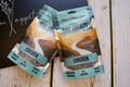 Two packages of chicken cat treats on a wooden surface with a dark background.|Deux paquets de friandises au poulet pour chats sur une surface en bois, sur fond sombre.