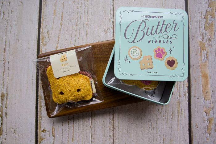 A cat toy duet cookie on a wooden tray next to its mint green tin box labeled Butter Nibbles.|Un jouet pour chat en forme de biscuit duo sur un plateau en bois à côté de sa boîte en métal vert menthe portant l'inscription Butter Nibbles.
