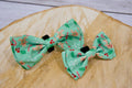Two green dog bow ties with Christmas festive patterns on a wooden surface.|Deux nœuds papillon verts pour chien à motifs festifs de Noël sur une surface en bois.