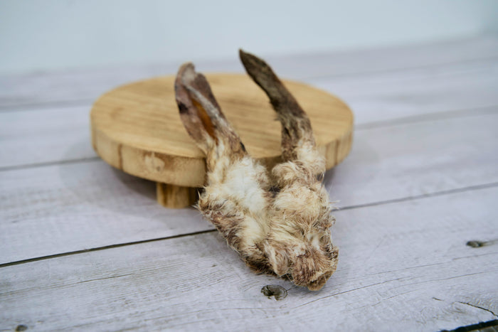 A pair of dried rabbit ears for dogs with attached fur, displayed on a round wooden stand.|Une paire d'oreilles de lapin séchées pour chiens, avec les poils, présentée sur un support rond en bois.