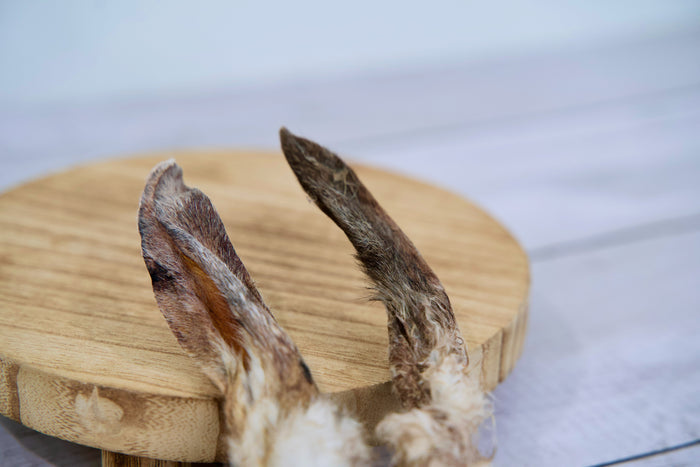 Two dried rabbit ears dog treats with natural fur, displayed on a round wooden board.|Deux friandises pour chiens à base d'oreilles de lapin séchées avec leur fourrure naturelle, présentées sur une planche en bois ronde.