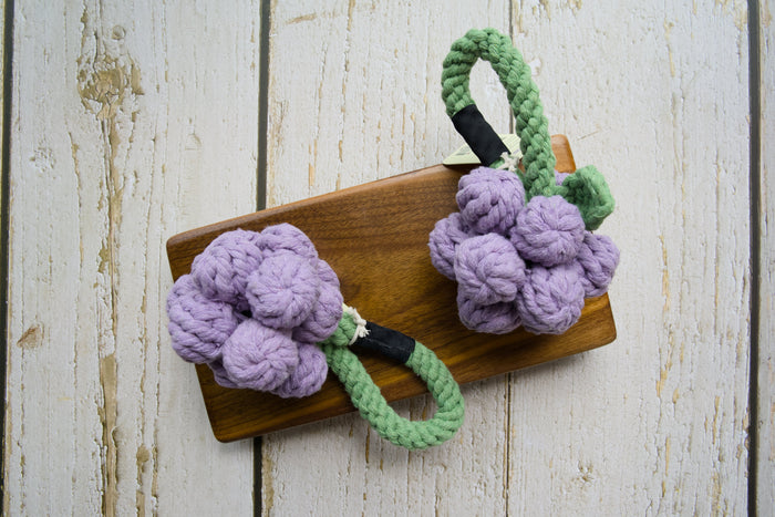 Two woven cotton grape clusters with green rope stems for dogs on a wooden board.|Deux grappes de raisin en coton tressé avec des tiges vertes en corde pour chiens, présentées sur une planche en bois.