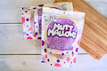 Packets of Mutt Mallows maple bacon kisses for dogs on a wooden surface.|Des sachets de friandises Mutt Mallows au bacon et à l'érable pour chiens, posés sur une surface en bois.