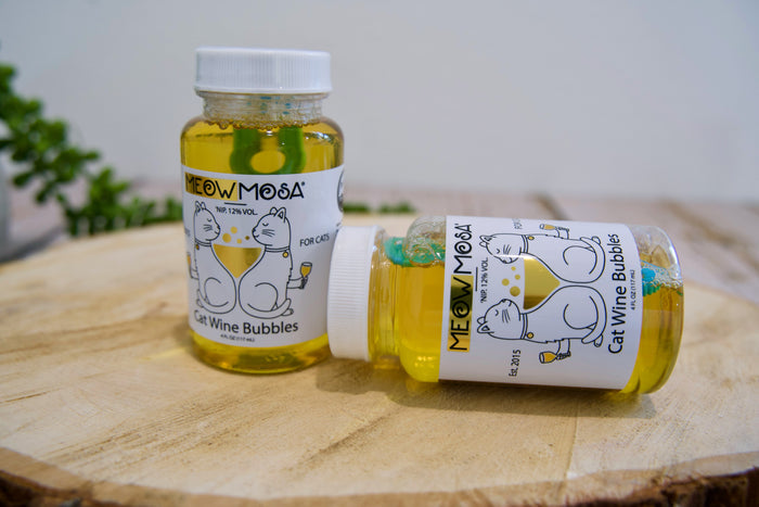 Two bottles of 'Meow Mosa' cat wine bubbles on a wooden surface with a blurred background.|Deux bouteilles de vin pétillant pour chats « Mew & Moos » posées sur une surface en bois, avec un arrière-plan flou.