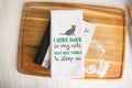 Cat-themed kitchen white towel with text and cat graphic on a wooden board.|Torchon de cuisine blanc sur le thème des chats, avec texte et motif de chat, posé sur une planche en bois.