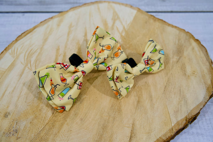 Two pet bow ties with colorful drink patterns on a wooden surface.|Deux nœuds papillon pour animaux à motifs de boissons colorées, posé sur une surface en bois.
