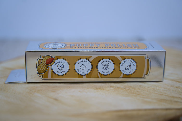 Box of a dog toothpaste with branding and ingredient information on a wooden surface.|Boîte de dentifrice pour chien avec la marque et les informations sur les ingrédients, posée sur une surface en bois.