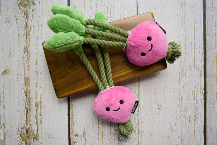 Two pink plush carrot-shaped dog toys with green leaves and rope tails, displayed on a wooden board.|Deux jouets pour chiens roses en peluche en forme de carotte, avec des feuilles vertes et des queues en corde, présentés sur une planche en bois.