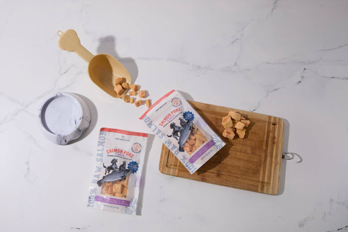 Two packages of salmon-flavored dog and cat treats are displayed on a white marble surface.|Deux paquets de friandises au saumon pour chiens et chats sont disposés sur une surface en marbre blanc.