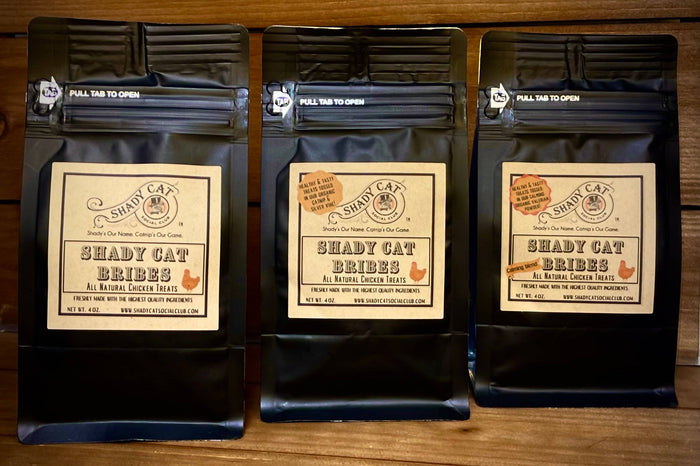 Three packages of Shady Cat Bribes cat treats on a wooden surface.|Trois paquets de friandises pour chats Shady Cat Bribes posés sur une surface en bois.