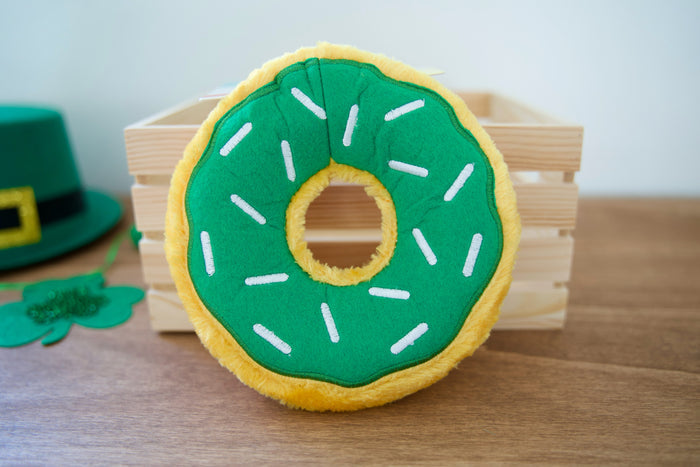 Green and yellow donut-shaped plush dog toy with a St. Patrick's Day hat in the background.|Jouet pour chien en peluche en forme de donut vert et jaune avec un chapeau de la Saint-Patrick en arrière-plan.