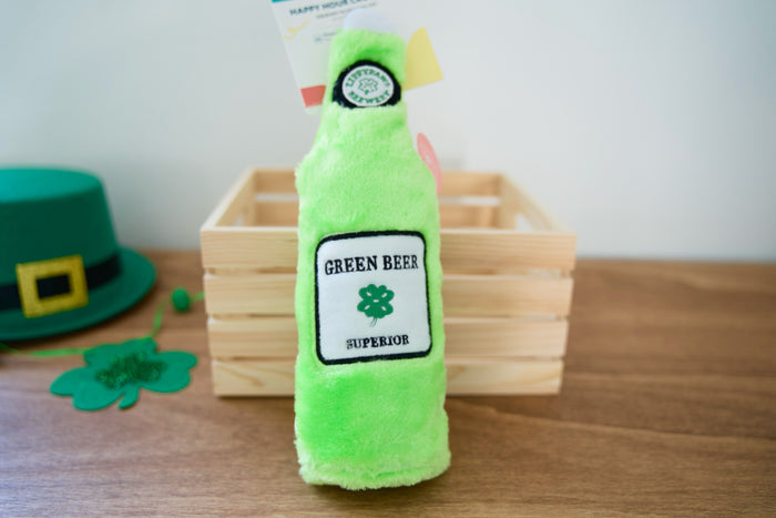 Green plush bottle dog toy with Green Beer Superior label on a wooden surface.|Jouet pour chien en peluche verte en forme de bouteille avec une étiquette Green Beer Superior sur une surface en bois.