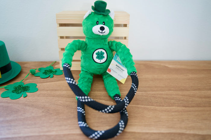 Green teddy bear with a top hat and shamrock emblem on a wooden table with a rope dog toy.|Un ours en peluche vert, coiffé d'un haut-de-forme et orné d'un trèfle, est posé sur une table en bois avec un jouet pour chien en corde.