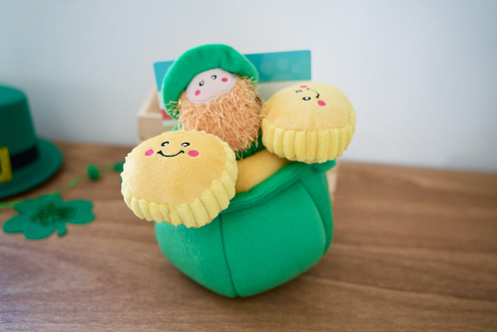 Plush toy for dogs with a leprechain in a green pot of gold on a wooden surface.|Peluche pour chien représentant un pot d'or avec un lutin sortant du pot posé sur une surface en bois.