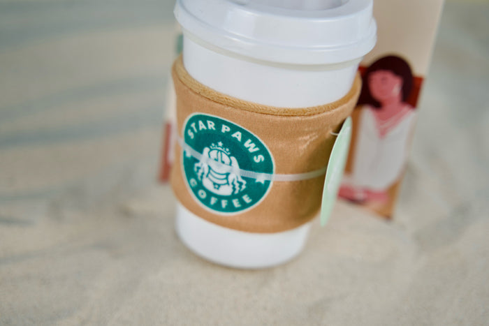 Vanilla-scented dog toy on sand, featuring rubber cup and plush sleeve for summer fun.|Jouet pour chien parfumé à la vanille sur le sable, avec tasse en caoutchouc et housse en peluche pour des jeux estivaux.