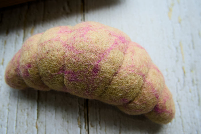 A handcrafted wool croissant dog toy with a beige body and pink swirls, resting on a white wooden surface.|Un jouet pour chien en forme de croissant, fabriqué à la main en laine, avec un corps beige et des spirales roses, posé sur une surface en bois blanc.