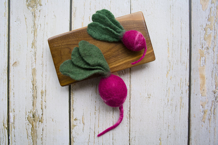 Two felted dog and cat toys shaped like radish with green tops and pink roots on a wooden board.|Deux jouets feutrés pour chiens et chats en forme de radis, avec des fanes vertes et une racine rose posé sur une planche en bois.