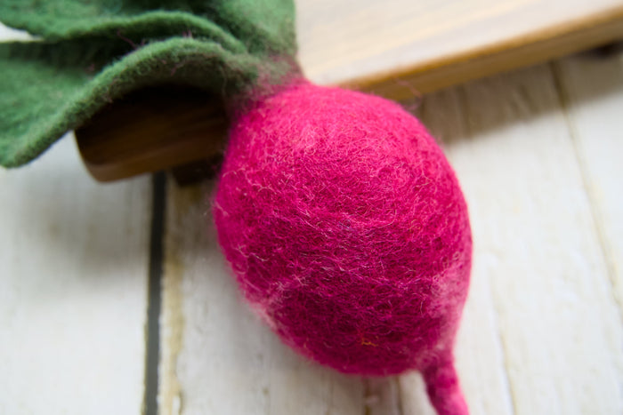 A close-up of a felted radish with a bright pink body and green felted leaves, resting on a white wooden surface.|Gros plan sur un radis en feutrine au corps rose vif et aux feuilles vertes en feutrine, posé sur une surface en bois blanc.