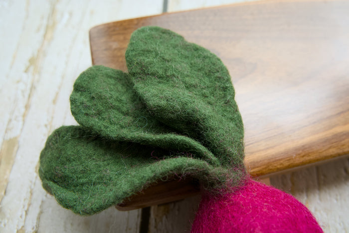 A close-up of a felted radish with green leaves and a bright pink root, resting on a wooden board.|Gros plan sur un radis en feutrine aux feuilles vertes et à la racine rose vif, posé sur une planche en bois.